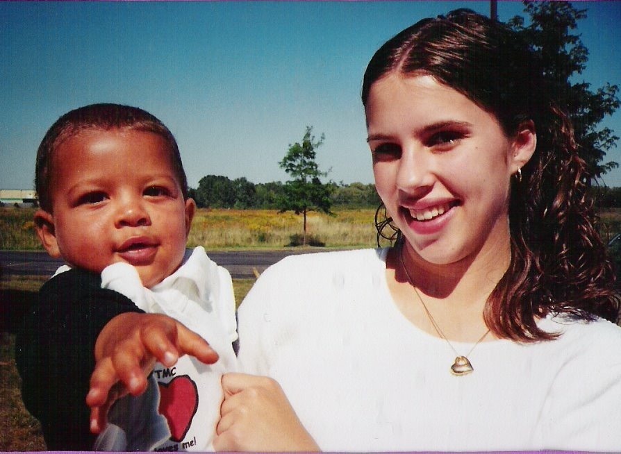 Young woman smiles while holding a toddler outdoors. The child reaches out playfully, both appearing joyful against a sunny landscape.