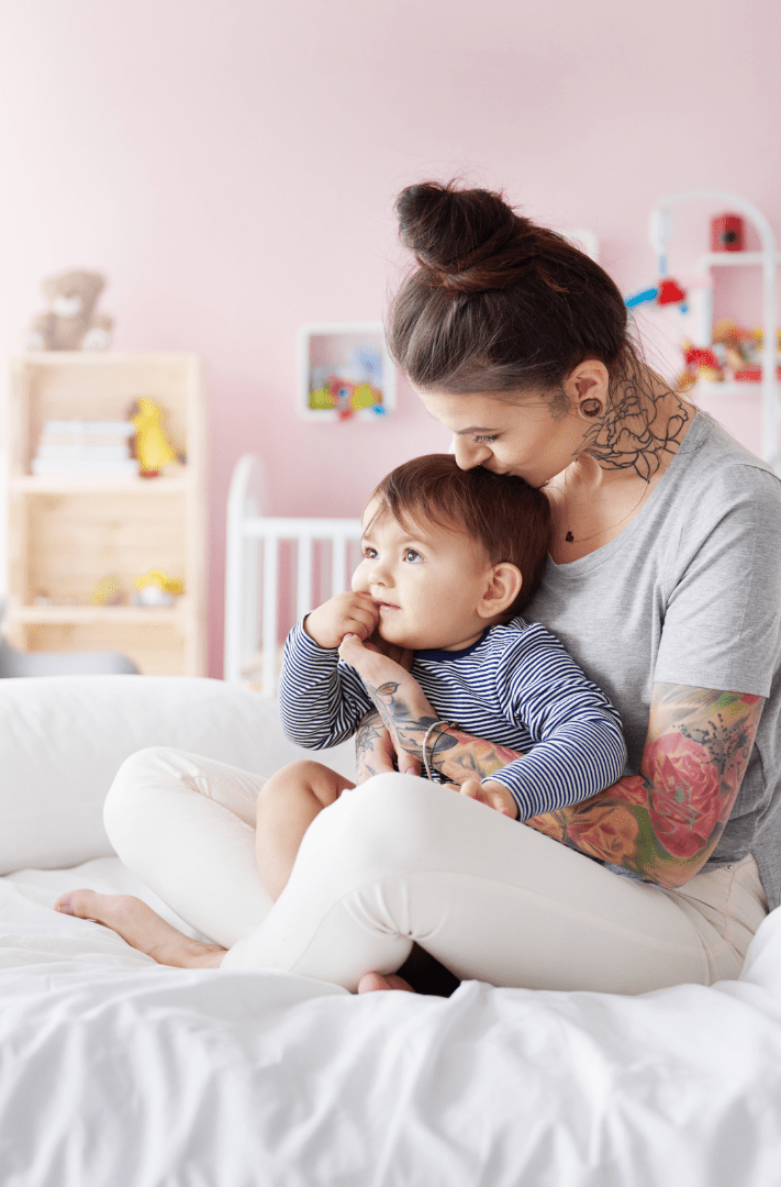 A mother with tattoos gently hugs her young child on a bed in a warmly decorated room, conveying a sense of love and intimacy.