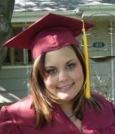 Smiling graduate in a maroon cap and gown, standing outside a residence. Celebrates a significant educational achievement.