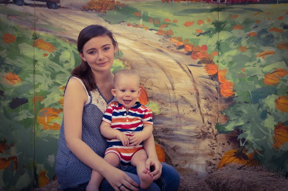 A young woman sits on hay, holding a smiling baby boy, against a colorful painted backdrop of a pumpkin patch. The image captures a moment of joy and connection.