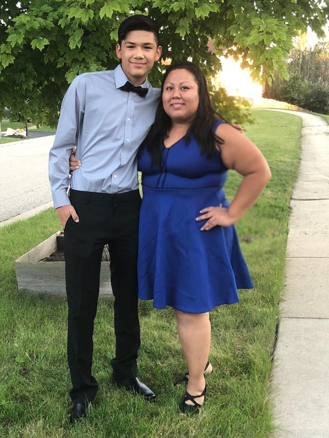 A young man in a light blue shirt and bow tie stands beside a woman wearing a blue dress, posing together outdoors.