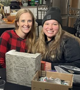 Two women smile together at a table decorated for a gathering, with a box of mugs and other gifts in front of them, capturing a joyful moment.