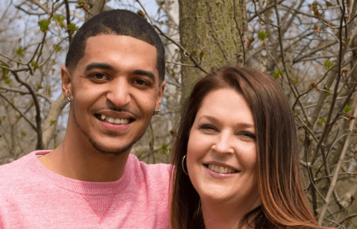 A young man and a woman smile together outdoors, surrounded by budding trees, conveying a sense of friendship or warmth.